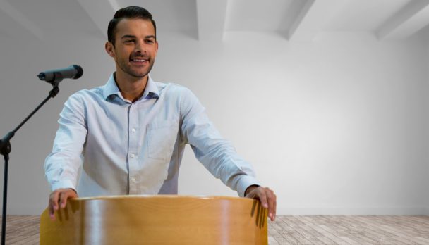 Businessman,On,Podium,Speaking,At,Conference,In,Room